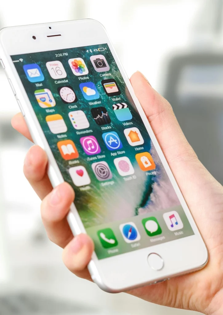 Eine Nahaufnahme einer Hand, die ein elegantes weißes iPhone schräg hält. Es präsentiert die komplette App-Suite mit Symbolen wie Mail, Kalender, Fotos und Kamera. Auf dem Display ist 14:36 Uhr vor einem leicht verschwommenen Innenhintergrund zu sehen.