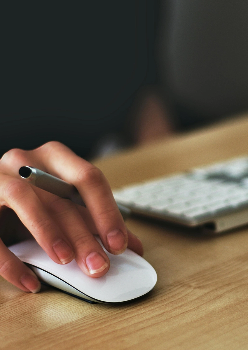 Eine Hand mit einer weißen Computermaus und einem Stift zwischen den Fingern liegt auf einem Holzschreibtisch und vermittelt die Atmosphäre eines voll ausgestatteten Arbeitsplatzes. Im Hintergrund ist eine unscharfe Tastatur zu sehen. Die Nägel der Person sind gepflegt, und die sanfte Beleuchtung verstärkt die ruhige Atmosphäre.
