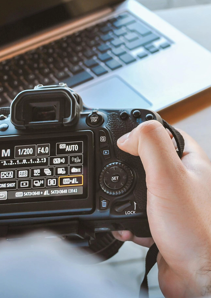 Eine Person nimmt Einstellungen an einer DSLR-Kamera vor. Auf dem Bildschirm werden Aufnahmeoptionen wie ISO, Verschlusszeit und Blende angezeigt. Die Kamera wird mit beiden Händen gehalten. Im Hintergrund ist auf einer Holzoberfläche ein Laptop zu sehen.
