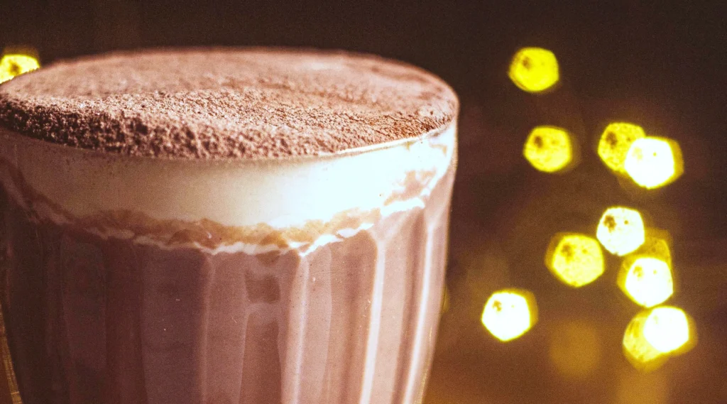Eine Nahaufnahme einer schaumigen heißen Schokolade mit Kakaopulver in einer Glastasse. Die Oberfläche ist cremig und samtig und erinnert an Mokka-Mousse. Im unscharfen Hintergrund erzeugen warme, gelbe Bokeh-Lichter eine gemütliche, festliche Atmosphäre.