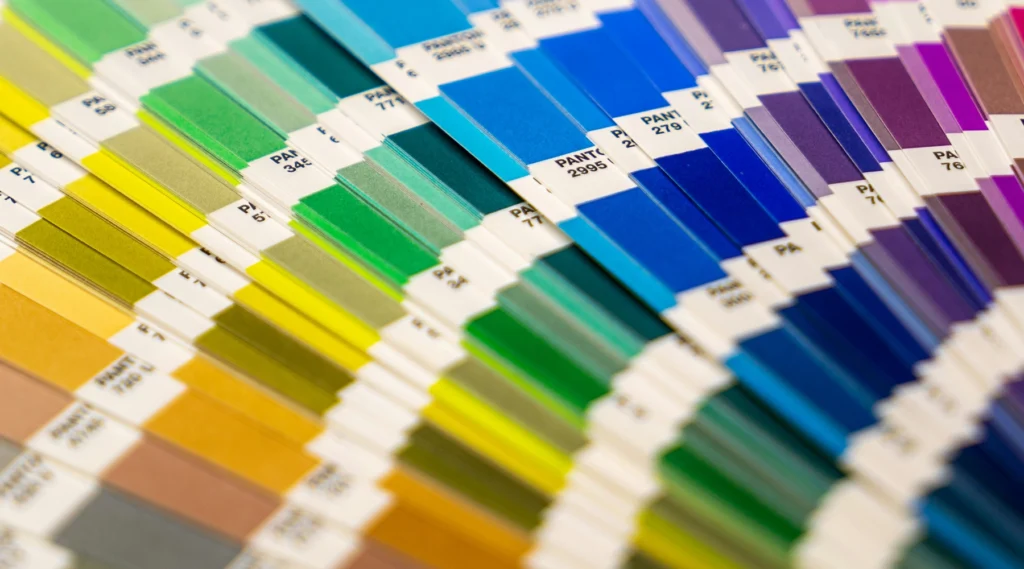 Nahaufnahme eines aufgefächerten Pantone-Farbmusterbuchs mit verschiedenen Farbtönen, die für jeden Entwurf unerlässlich sind. Die Farbmuster wechseln von Grün und Gelb links zu Blau und Lila rechts, jeweils mit Pantone-Nummern. Kräftige Farbtöne sind zur leichteren Übersicht in überlappenden Reihen angeordnet.