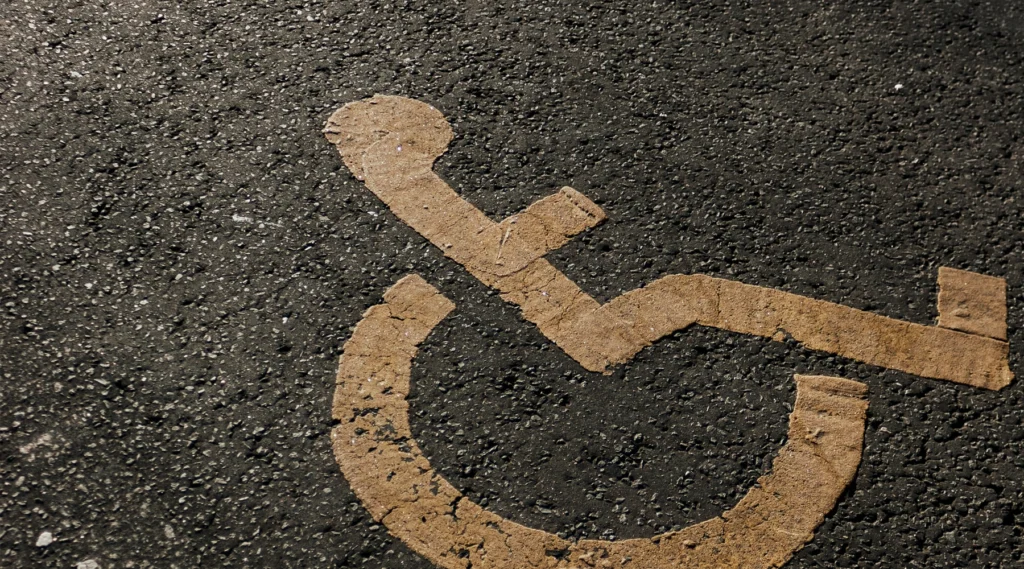 Ein verblasstes gelbes Rollstuhlsymbol auf strukturiertem Asphalt markiert einen barrierefreien Parkplatz. Die abgenutzte, abgeplatzte und abblätternde Farbe verschmilzt mit der dunklen, mit kleinen Steinen übersäten Straße und symbolisiert die Notwendigkeit kontinuierlicher Verbesserungen der Benutzerfreundlichkeit, um eine wirklich barrierefreie Website zu schaffen.